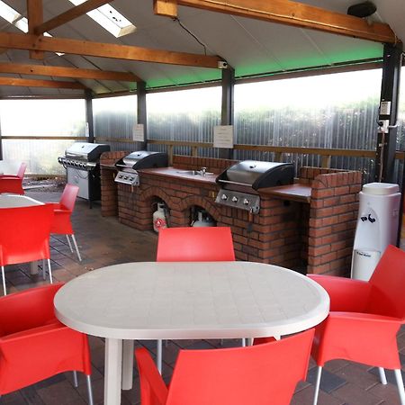 陶波德布雷思汽车旅馆 外观 照片 The photo shows an outdoor kitchen area with a covered space. There are several barbecue grills set up on a brick counter. In the foreground, there are round tables with white tops and bright red chairs arranged around them. A Coca-Cola vending machi