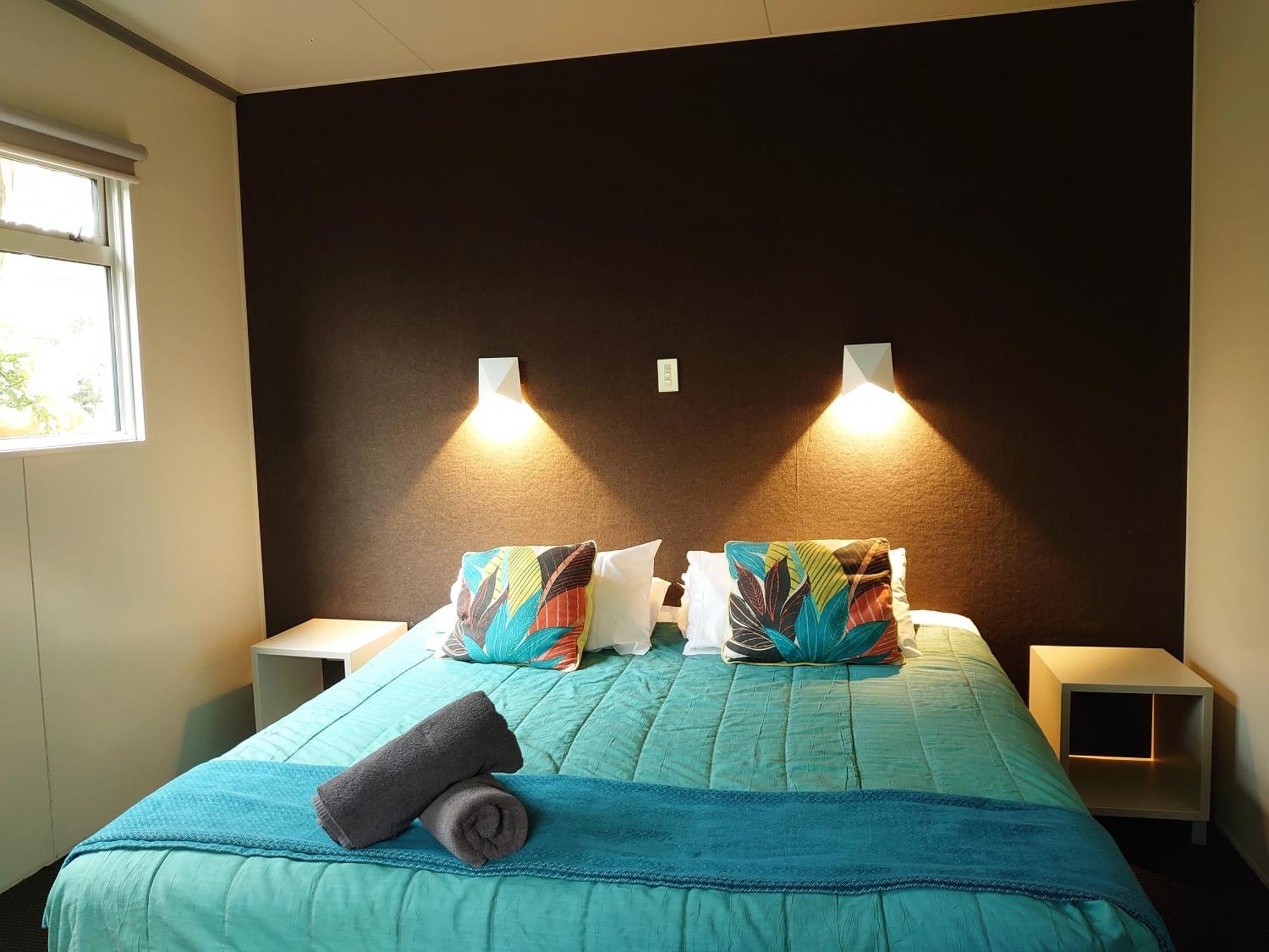 陶波德布雷思汽车旅馆 外观 照片 The image shows a bedroom with a modern and inviting layout. The focal point is a bed with a vibrant turquoise comforter, decorated with two colorful pillows featuring floral patterns. On either side of the bed, there are small nightstands with wall-