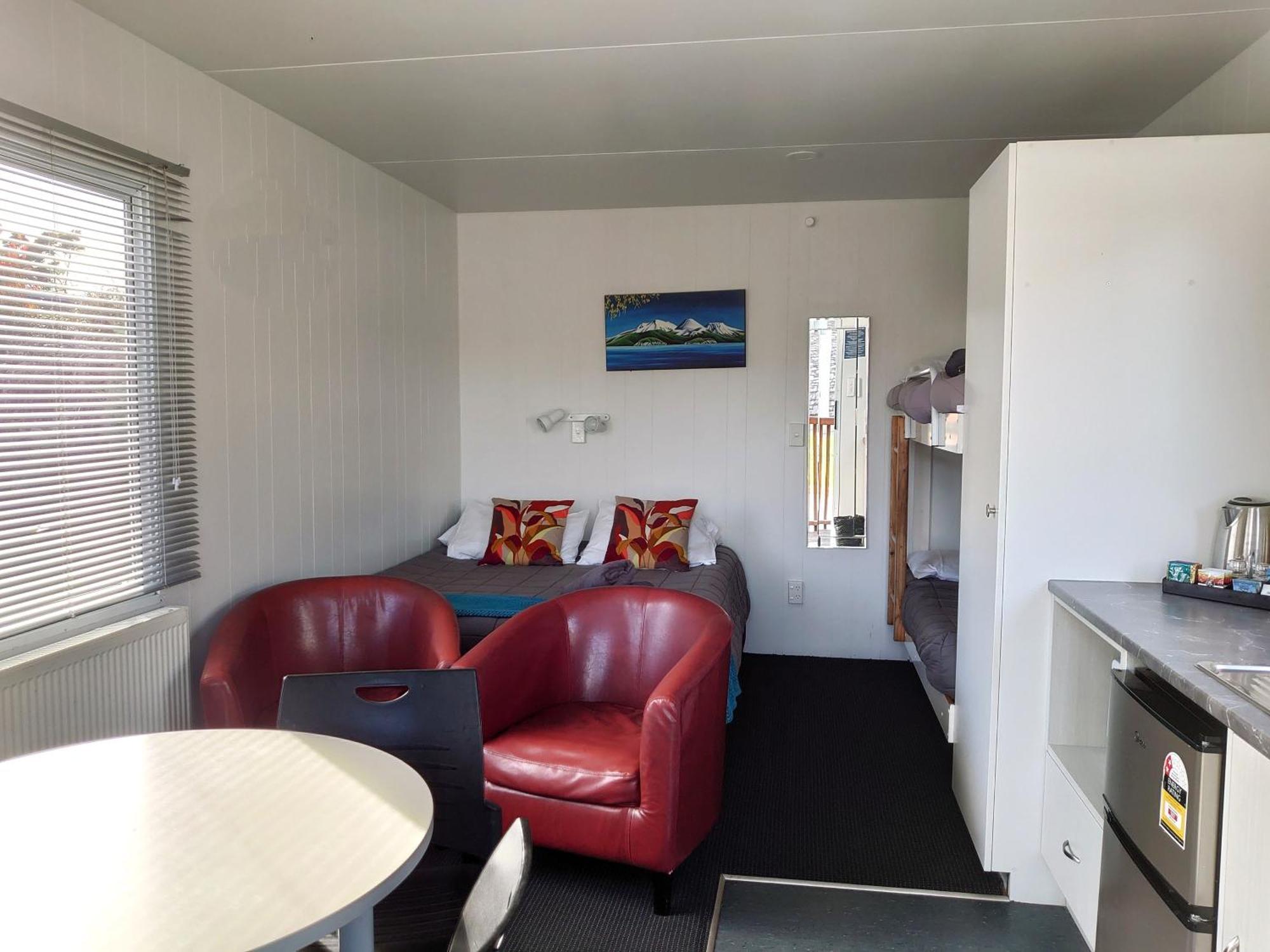 陶波德布雷思汽车旅馆 外观 照片 The photo shows the interior of a small living space, possibly a cabin or studio apartment. There are two red armchairs and a round table in the foreground. The wall opposite has a bed with colorful pillows and a small nightstand. To the side, there 