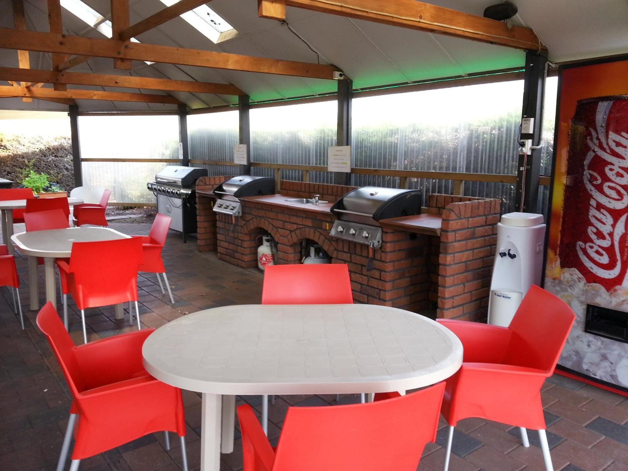 陶波德布雷思汽车旅馆 外观 照片 The photo shows an outdoor kitchen area with a covered space. There are several barbecue grills set up on a brick counter. In the foreground, there are round tables with white tops and bright red chairs arranged around them. A Coca-Cola vending machi